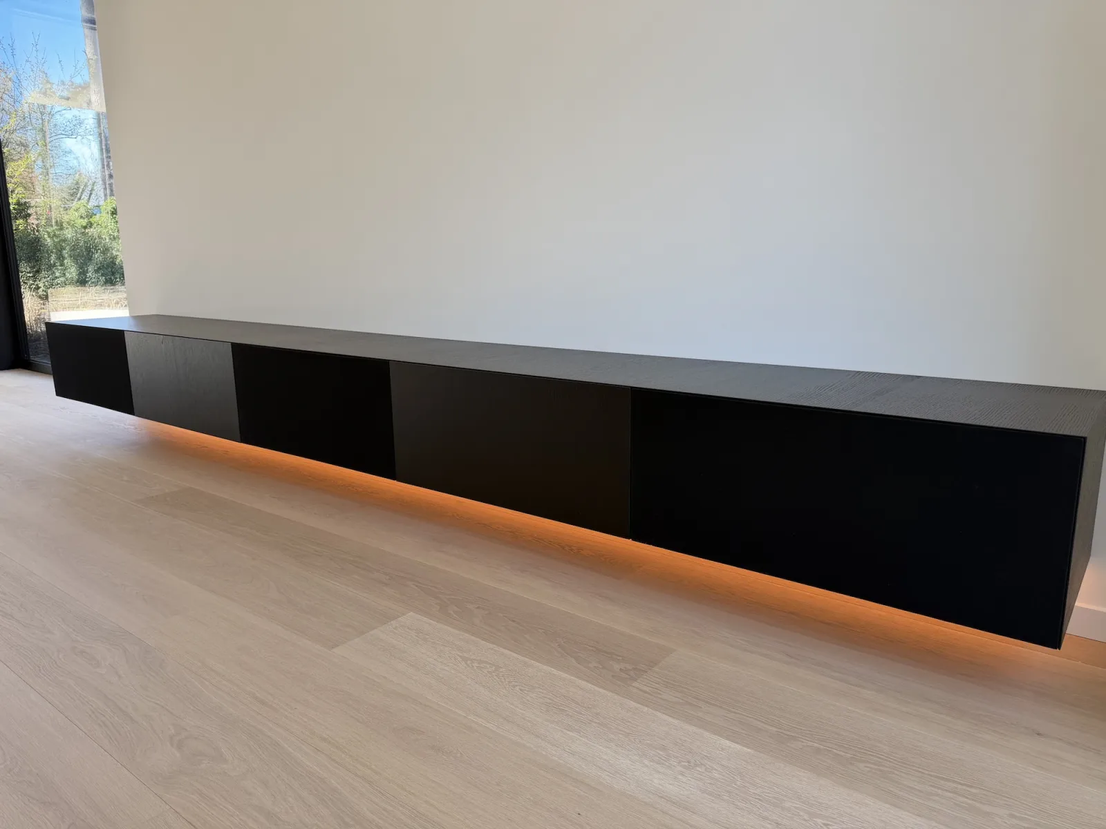 Lang zwart zwevend maatwerk tv meubel met een oranje ledlijn onder de bodem, naast een raam tot de vloer